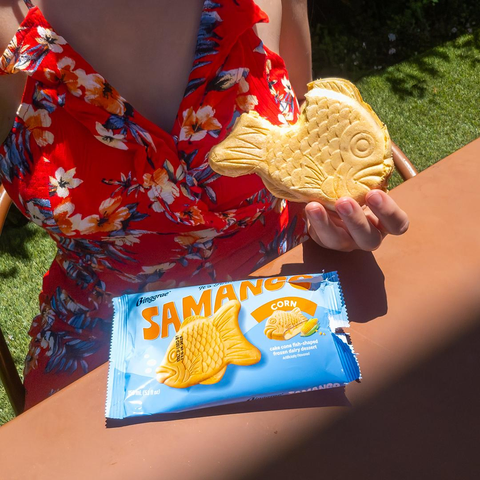 Photo of Lety sitting at an outdoor dining table holding a fish‐shaped, corn‐flavored ice cream sandwich with a big bite taken out of it during high noon. From top to bottom, her visible attire consists of: a floral red sundress. Lety is sitting on a brown metal chair at a matching outdoor dining table, holding her phone in her right hand to take a top‐down selfie that focuses on her torso. In her left hand is a corn‐flavored store‐bought samanco, the wrapper of her frozen dessert on the table in front of her, and green artificial turf around the floor behind her. Lety's late, former, ex‐fiancée… she'll never find the right words to describe their relationship, but someone Lety loved very much loved corn‐flavored everything. She's still trying to adjust to her lover's palette, three years after she's gone. A basic transcript of the samanco wrapper follows.

Binggrae®
It's Fintastic!
SAMANCO
CORN
cake cone fish‐shaped frozen dairy dessert
Artificially Flavored
150 mL (5.1 fl oz)