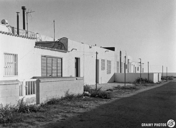 Black and white photo of a row of simple, flat-roofed houses along a dirt road. The scene includes a low wall, plants, and utility poles, conveying a quiet, rural atmosphere.