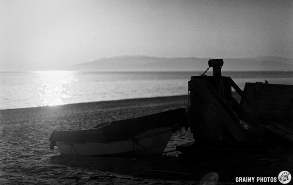 A serene beach scene in black and white, featuring a small boat resting on the sand alongside a weathered wooden structure, with soft waves and distant mountains illuminated by the sunlight reflecting on the calm Mediteranean sea.