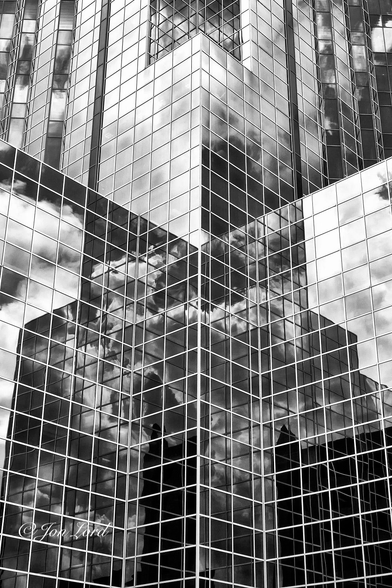 This is an abstract architectural photo in portrait format of the lower floors of a high-rise office block with numerous faceted facades made up multiple panes of glass that reflect the clouds surrounding the building. Houston, Texas (2012).

The camera is located about ten or more metres away and angled towards the centre of one of the facets comprising of a concave 'V' shape. From the centre, two tall walls of glass radiate outwards to the left and right edges of the photo. This part of the building has 15 floors in view., with some more out of view below the photo. Above, the facade reverses with a sharp edged, right angled corner in the centre of the image and the walls of glass viewed as a convex 'V' shape (this, though unseen, is the main square shaped, but tall tower block rising 64 floors). The entire walls are made up of uniform squares of glass set in silver frames. The glass, while reflecting a blue sky, are rendered black with the white fair weather cumulus clouds reflected in most of the panes of glass.