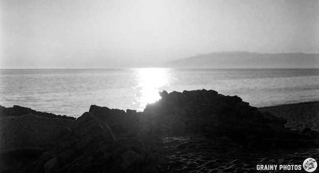 A serene black and white beach scene, featuring rocky formations in the foreground, with the shimmering sun reflecting on a calm sea. The distant shoreline fades into soft haze, creating a tranquil atmosphere.