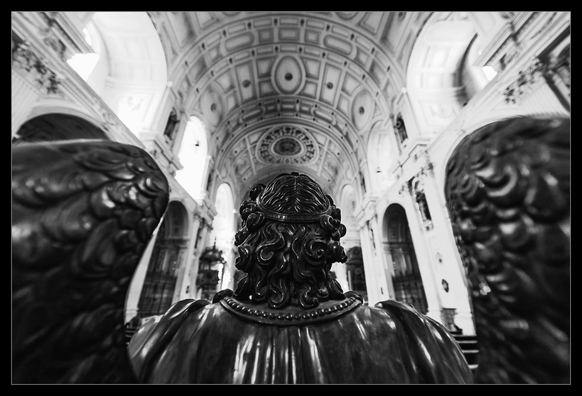Eine menschengroße Statue eines geflügelten Engels in einer Kirche. Die Aufnahme ist mit einem Ultraweitwinkel von der Rückseite der Statue  angefertigt worden mit fokus auf dem Hinterkopf. Flügel und die Kirche im Hintergrund sind aufgrund Tiefenunschärfe ... naja ... unscharf.