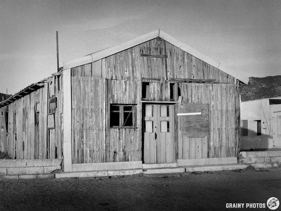 A weathered wooden house with a peaked roof, featuring two front doors and multiple windows. The structure shows signs of wear and is set against a barren landscape, captured in black and white.