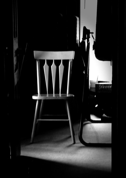 A white wooden chair sits in the shadows of a small room. The light from a window on the left and a window behind are seen in this high contrast black and white image.