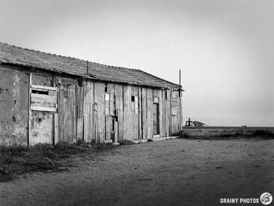 Black and white photograph of weathered wooden structures along a sandy path, with a serene sea view in the distance. The buildings display rustic charm, featuring various textures and details from years of exposure to the elements.