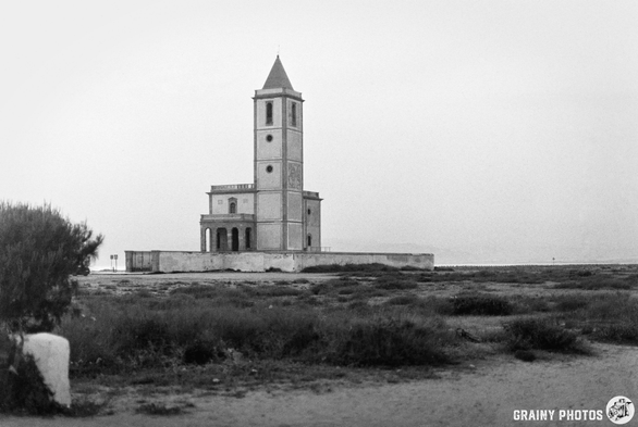 The solitary tower of Las Salinas church stands on a sandy landscape, surrounded by sparse vegetation. The church features a peaked roof on the bell tower and multiple windows, with calm waters visible in the background under a hazy sky.