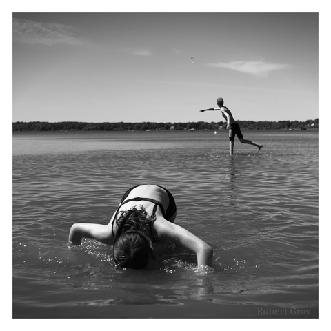 Monochrome image of a girl dipping her face in the water. In the background and further from the shore is a boy wearing trunks who is throwing an object away from the camera.