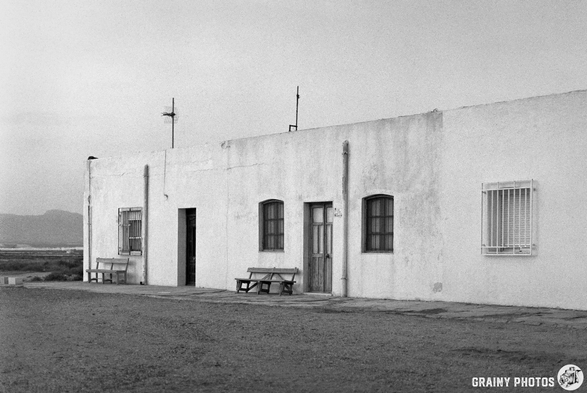 A rustic, white building with a gravel foreground, featuring multiple doors and windows. Several benches are placed in front, creating a simple, tranquil ambiance against a backdrop of distant hills. The image is in black and white, enhancing its nostalgic feel.