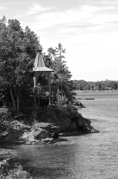 A small ornate gazebo is seen perched atop the rocky shoreline of a large lake. Large trees surround it and the shoreline stretches into the background and to the right. Some clouds fill the sky on this summer day and the water is calm. This is a black and white image.