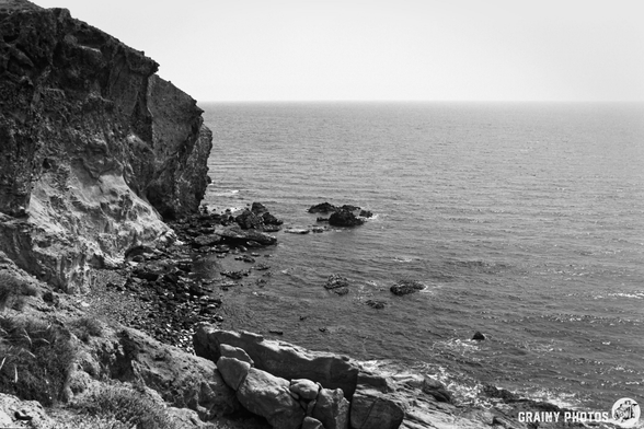 A black and white coastal scene featuring rugged cliffs beside a calm sea, with scattered rocks along the shoreline and a serene horizon.