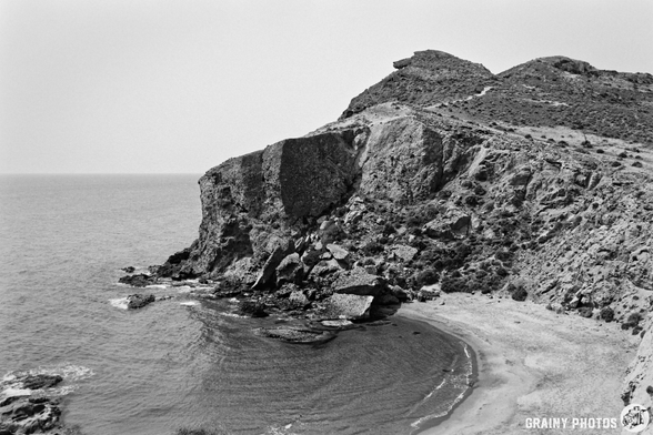 A dramatic black-and-white scene showcasing a rugged coastal cliff overlooking a calm sea, with rocky formations and a curved sandy beach. The landscape captures the serene beauty of nature, emphasizing textures and contrasts.