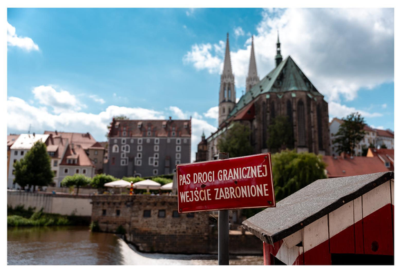 Blick über einen Grenzfluss auf eine Altstadt mit Kirche. Im Vordergrund ist noch ein Grenzhäuschen (aus Zeiten des kalten Kriegs) zu sehen. Ein Schild auf Polnisch informiert: Grenzstreifen – Betreten verboten. 