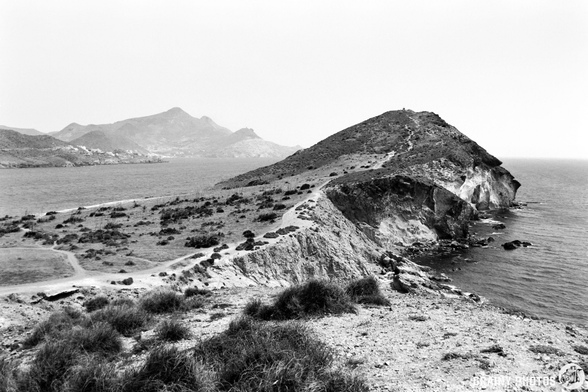 A rocky coastline with rugged cliffs and a winding path leads to a distant mountain range. The scene is captured in black and white, highlighting the dramatic landscape and tranquil sea on a cloudy day.