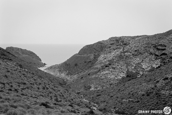 A monochrome landscape featuring a valley between rocky hills that leads to a calm sea in the distance, with a hazy sky above and sparse vegetation on the slopes.
