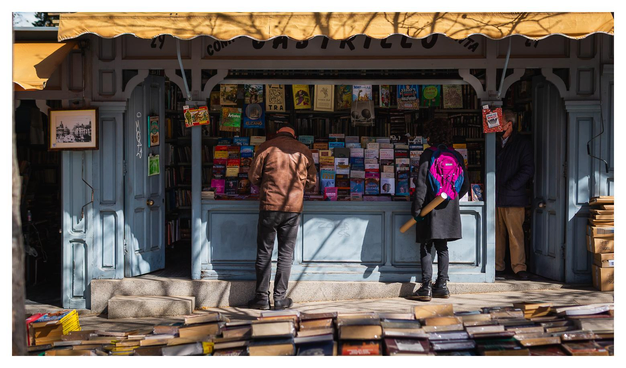 Eine Art Kiosk bzw. Bude aus dem/der gebrauchte Bücher direkt auf die Straße verkauft werden. Im Vordergrund ist ein Tisch zu erkennen, auf dem ebenso Bücher liegen. Mehrere Menschen stehen herum und suchen nach Lektüre.