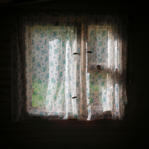 This color photograph captures a window in an old wooden house. The room is immersed in semi-darkness, but the window is brightly illuminated by sunlight from outside. Light, translucent curtains with a delicate blue floral pattern hang on the window. The light filters through the thin fabric, illuminating it from within and creating a soft, diffused glow. Green garden leaves and sunbeams can barely be seen through the curtains. The dark wooden walls and window frame create a deep contrast, highlighting the glowing rectangle of the window. The image conveys a sense of peace, coziness, and quiet, sunny summer life.