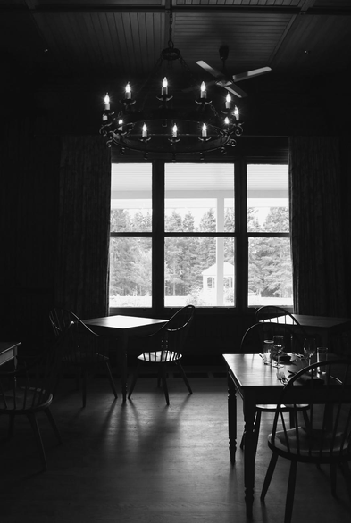 Black and white photograph of a dark dining room in an old house, lit by a circular chandelier of electric candles hanging from the ceiling of wooden slats, and natural light coming from a large window. Natural light is reflected on the wooden surface of three tables and their two chairs each, and on the wooden floor. Two cutlery sets with glasses and cups are placed on one of the tables. A landscape of tall trees with a pavilion on the lawn is visible through the window.
Photographie noir et blanc d'une salle à manger sombre dans une maison ancienne, éclairée d'un lustre circulaire de chandelles électriques accroché au plafond de lattes en bois, et de la lumière naturelle venant d'une grande fenêtre. La lumière naturelle se reflète sur la surface en bois de trois tables et de leurs deux chaises chacune, et sur le plancher de bois. Deux couverts avec des verres et des coupes sont placés sur une des tables. Un paysage de grands arbres avec un pavillon sur la pelouse est visible par la fenêtre.