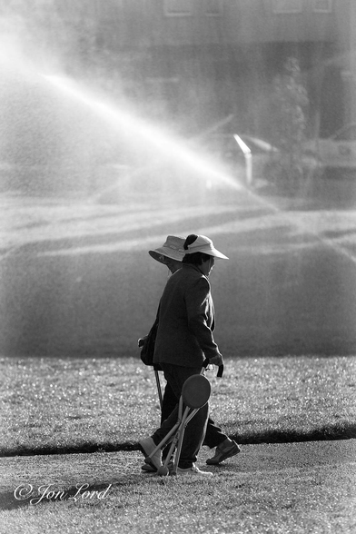 This is a black and white street photo in portrait format of two older Chinese ladies passing each other on a park footpath with two jets of water forming an 'X' over the ladies with much of the background obscured in a mist of spray. Washington Square, San Francisco (2013).

In the immediate foreground is a patch of light grey toned grass with a footpath beyond that stretches across from left to right. On the far side of the footpath is more grass. The first part, beyond, rising up to the quarter up level, is lit by the morning sunlight. Beyond, the grass park rising up to the three-quarters up level is deep in shadow. In the centre of the photo are our two Chinese ladies, viewed from a few metres away, are walking in opposite directions and passing each other, shoulder-to-shoulder. The closer lady is walking from left to right, is wearing a white cap with a large visor, a dark fleece, dark trousers and white trainers. She is carrying a walking aid in her right hand that doubles as a walking stick and stool. Partially obscured by the first lady is our second Chinese lady, walking from right to left, wearing a large straw hat and dressed in a similar style to the first lady other than having a simple walking stick. Beyond our two ladies are the two strong jets of water, both angled upwards at about forty-five degrees and forming a 'X' as the two jets cross above the park. Surrounding this scene in the background is a misty cloud of water spray from the irrigating jets of water. Barely visible in the background and through the mist is a distant street scene, about ten metres away, with a few cars, trees and buildings evident. 
