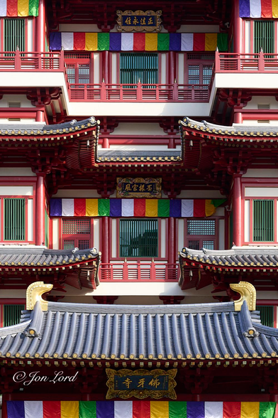This is a colour photo in portrait format of the front facade of a multi-floored, highly ornate and colourful, Buddhist temple located in Chinatown, Singapore (2012).

Extending along the base of the photo is a string of multi-coloured, square flags that stretch from the left to the right edge of the photo, this is above the entrance which is out of view below. Above is a tiled, pitched roof that again stretches across the image. The roof is covered with glazed, grey, oriental tiles with a lintel that extends along the ridge of the roof with a large bright golden mythical figure head at each end of the ridge. Between the roof and the row of flags is a large rectangular plaque with a golden filagree surround and three large golden Chinese characters. The windows of the first floor are mostly obscured by the entrance roof. Above the first floor windows is a small roof leaning out in front of the building, one either side of the centre. These small roofs are again made from grey, glazed Chinese tiles. The second floor windows are clearly in view. The windows are a mix of vertical, rectangular and square shapes, all with a red wooden surrounds and with vertical open, green blinds inside. Between the two small overhanging roofs is a large central square window with a line of fifteen rectangular multi-coloured flags above, and a further ornate plaque with four golden Chinese characters. Above might be the third floor which is obscured by the small roofs. Above is the top floor which has a balcony with a red wooden railing at the front with a further string of flags stretching the entire width of the building and topped with one last plaque with four Chinese characters. 



