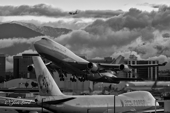 This is a black and white photo in landscape format of a scene at a busy airport with a Boeing 747 taxying in the foreground, a further 747 that's just taken-off, clouds and mountains in the background and a smaller aircraft on it's approach. LAX, California (2018).

Stretching along the entire base of the photo is our taxying 747, taxying from left to right. The plane is viewed from slightly above, at a distance of 50m away and with the tail and right side in view. On the tail is a large logo of a plum blossom, while just aft of the nose are the words 'China Airlines' and 'CARGO' in larger lettering (Republic of China (Taiwan)). This aircraft is easily identified as a freighter since it has no passenger windows. Above the lettering is the distinctive hump that the 747 has for the upper deck and flight deck. A short distance beyond is the further 747 that has just taken-off. It is in the centre of the photo, above the first 747 and in-flight from right to left and about 100m from the first aircraft. This 747 has it's nose pointing skywards, it's undercarriage is still down and streams of heat haze can be seen extending behind the engines. This 747 has a longer upper deck stretching back to the leading edge of the wings with the words 'Asiana Airlines' just below the upper deck. In the middle background area is a row of medium-rise office and hotel buildings spreading from the left to the right side of the photo. Above and behind the buildings is a mass of low clouds sloping downwards from the upper right corner towards the middle of the left edge of the image. In the gap to the upper left side is the approaching aircraft, about 1km away, its gear and flaps down and landing lights on. In the far distance, again in the upper left corner is a range of snow covered mountains. 