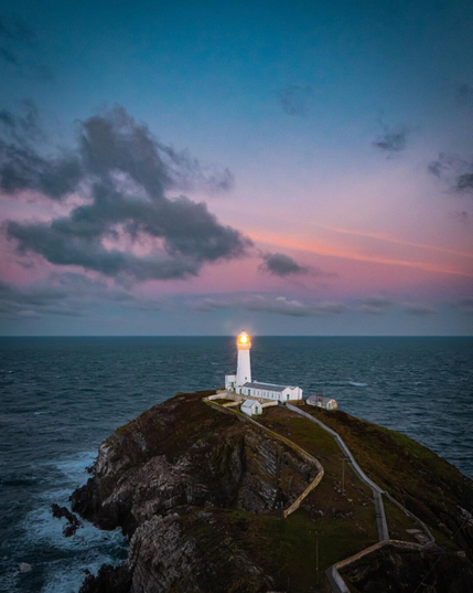 A high-angle photo of the white South Stack Lighthouse at sunrise. The beacon at the top of the tower glows with a bright orange light. The lighthouse and buildings sits on a rocky, dark green outcropping surrounded by choppy dark blue waters. A narrow path with a stone wall winds down the cliff towards the lighthouse buildings. The sky is soft blue, pink, and purple with dark, clouds.