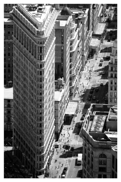 A view from above the Flatiron Building in Manhattan, New York City, on a bright, sunny day when the light casts strong shadows and the people walking on Fifth Avenue are tiny compared to the monolithic buildings.