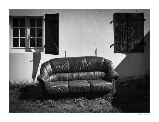 Eine schwarz-weiße Fotografie zeigt ein altes Ledersofa, das vor einer hellen Hauswand steht. Links befindet sich ein Fenster mit geöffnetem Fensterladen, rechts ein weiteres Fenster mit geschlossenem Fensterladen. Das Sofa wirft einen deutlichen Schatten auf den Boden, und Sonnenlicht fällt schräg auf die Szene.