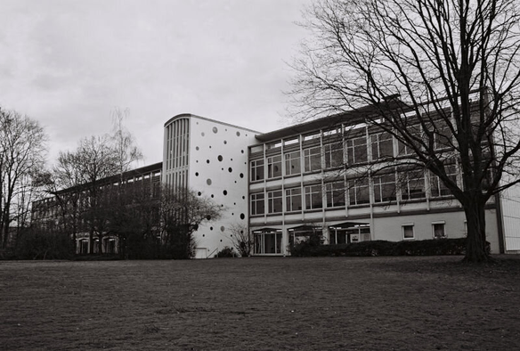 A modern building that is mostly plain boxes but there is a center section with large windows, a curved roof, and smaller round windows at varying sizes and intervals on the side.