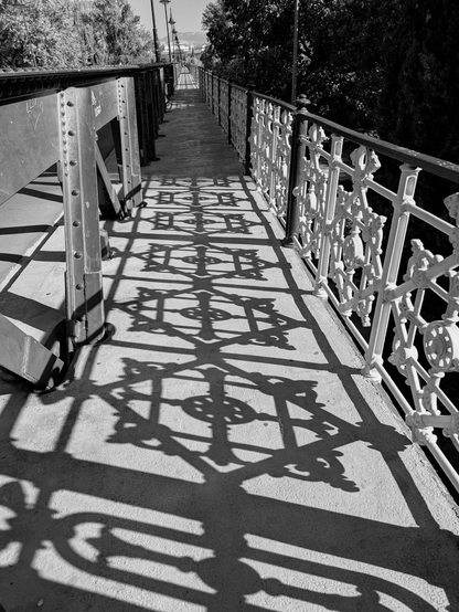 Black and white picture looking down the pavement portion on the right side of the road and bridge. Large iron beams and rivets are to the left and iron railing with pattern is to the right. The pavement between the beams and railing are filled with the shadows of the railing design.