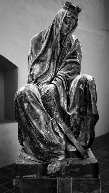 A black-and-white photograph of a seated bronze statue of a robed woman on a rectangular pedestal, representing the city of Magdeburg after the Sack of Magdeburg. She is draped in flowing robes covering her body and head, with only her hands and feet visible, her head bowed in a posture of grief. Her hands hold a sword in her lap. The setting is an indoor space with plain, light-coloured walls and a tiled floor.