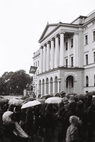 Black-and-white analogue photograph of the Norwegian Royal Palace. A crowd is gathered outside, carrying umbrellas and wearing raincoats. A long flag pole juts out from the balcony, with the royal standard flying at half mast, topped with a black mourning veil.