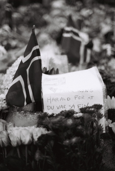 A black-and-white analogue close-up photograph of a sea of flowers and bouquets, dotted with small Norwegian flags. In the foreground lies a piece of paper in a rain-covered plastic pocket. The paper has a drawing of a Norwegian flag, a heart, and the text ‘Thank you King Harald for being so nice’ in a young child's handwriting.