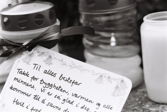 A black-and-white analogue close-up photograph of several grave lanterns. One has a card tied to it with a ribbon. The card is addressed ‘To everyone's grandfather’ in neat handwriting.