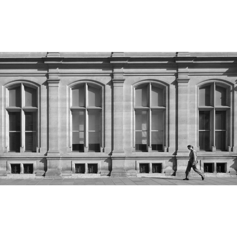 Black and white street photography taken in Paris showing a woman passing by the windows of a classical building. The four windows look very similar to each other.