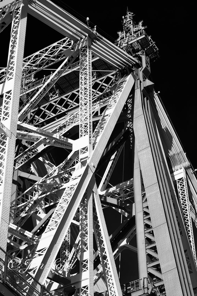 This is a high contrast abstract, black and white photo in portrait format of the tower section of a historic cantilever road bridge brightly lit by the still low morning sun with a clear and cloudless sky rendered jet black. 59th Street Bridge, NYC (2012).

The camera is angled steeply upwards at a section of the tower of this steel cantilever bridge. The white lattice and truss metal work is brightly lit by the just risen morning sun. The structure is an orderly jumble of straight lengths of steel, mostly trusses made up from metal formed into lattices and then riveted into a long box like trusses. Some of the lengths are vertical, some horizontal and a few diagonal. Just visible spanning the top are long lengths of steel plating, all of the lengths closely spaced together and extending from the anchorage, on land, over the towers to the far anchorage with vertical struts descending down and supporting the road deck below, which in this case is hidden below the base of the photo. 

