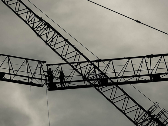 Silhouetted workers are seen on a construction crane against a cloudy sky, with intersecting crane beams creating a dynamic composition.