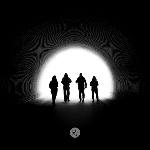 Ein Schwarz-Weiß-Foto zeigt die Silhouetten von vier Personen in einem Tunnel. Die Personen befinden sich in der Bildmitte und bewegen sich auf eine helle, weiße Öffnung am Ende eines dunklen, kreisförmigen Tunnels zu. Die Wände des Tunnels weisen eine gerippte Textur auf. Die Personen erscheinen als dunkle Umrisse vor dem starken Gegenlicht. Am unteren Bildrand befindet sich ein kleines, weißes kreisförmiges Logo mit einem stilisierten Symbol.