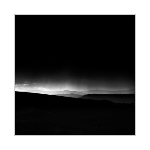 Square black-and-white photograph. A nearly black sky fills most of the upper frame, fading to a narrow band of light along the distant horizon. Several small wind turbines stand on a pale ridge left of centre. Overlapping hills become progressively lighter and less distinct in the distance, while a nearly black foreground slope descends from left to right across the lower portion. The light is strongest near the left horizon, where a few bright cloud edges are visible.