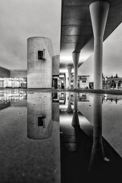 Eine Schwarz-Weiß-Fotografie zeigt eine markante, moderne Betonarchitektur bei bewölktem Himmel, die sich detailreich in einer großen Pfütze im Vordergrund spiegelt. Auf der rechten Seite erstreckt sich ein langes, erhöhtes Betondach, das von mehreren hohen, sich nach oben hin kelchförmig verbreiternden Säulen getragen wird. Links daneben steht ein massiver, zylindrischer Turm aus Sichtbeton, an dessen oberem Teil ein kleines, Fenster zu erkennen ist. Im Hintergrund schließen sich flache, verglaste Gebäudeteile an, in denen Licht brennt. Der Boden im Vordergrund ist nass, und die perfekt glatte Wasseroberfläche wirft ein scharfes Spiegelbild der gesamten Szenerie nach unten, wodurch eine Symmetrie zwischen der realen Architektur und ihrer Reflexion entsteht.