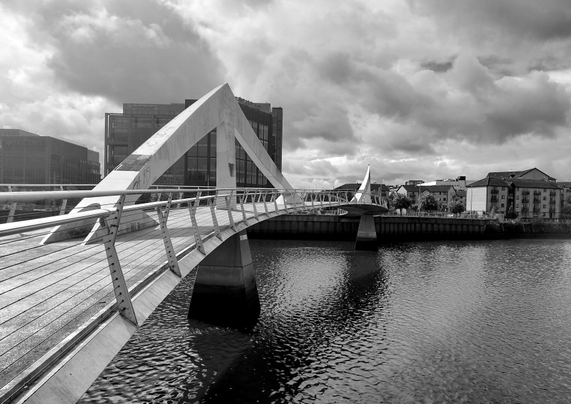 Black and white photo of Tradeston Bridge, Glasgow, an S shaped pedestrian bridge. A walkway with metal railings curves across the river, supported by a heavy concrete pier fixed into the riverbed. There's a prominent white triangular arch structure (a fin) in the middle, and a second matching arch in the distance, on the far side of the bridge. On the riverbank in the distance, modern multi-storey buildings can be seen under a moody, cloud-filled sky.
