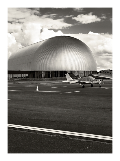 Im Vordergrund steht ein kleines Flugzeug auf dem Rollfeld, daneben ein Verkehrskegel. Im Hintergrund erhebt sich der Luftschiffhangar am Flughafen Mülheim/R., dessen gewölbte, metallisch wirkende Außenhaut die Szene dominiert. Die Aufnahme ist in schwarz-weiß gehalten, wodurch die Wolken am Himmel und die Struktur des Hangars besonders betont werden. Die Abmessungen des Hangars betragen L 92 m, W 42 m, H 26 m, Weight 557 t wood