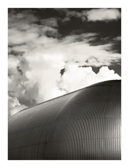 Schwarz-weißes Foto eines Luftschiffhangars mit gewölbter, gerippter Metallfassade im Vordergrund. Im Hintergrund sind große, dramatische Wolkenformationen zu sehen. Die Bildkomposition betont die Rundung und Struktur des Hangars im Kontrast zu den weichen Wolken.