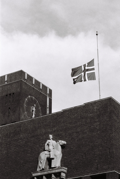 Black-and-white analogue photograph of Oslo City Hall, showing the West wall, a flag pole with the Norwegian flag at half-mast, and part of the city hall's clock tower opposite it. Flush against the large brick wall is a stone statue of King Harald III Hardrada sitting on a horse, sculpted by Anne Grimdalen in 1950. The dark brick building with its square shapes and geometric brickwork stands in contrast to the heavy but bright clouds above.