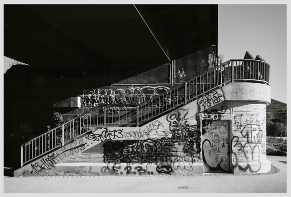 Horizontal black and white image of stairs leading up to a railroad bridge with a footpath. The concrete base of the bridge is covered in graffiti.