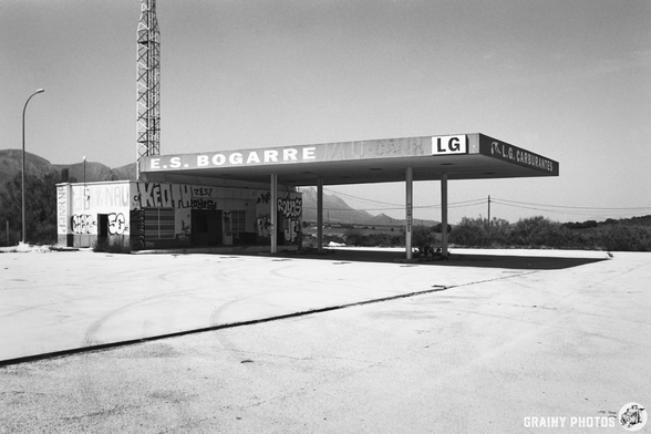 Abandoned petrol station with graffiti on the building and forecourt, surrounded by mountains and sparse landscape in a black and white setting.