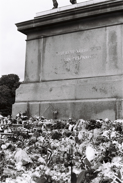 Black-and-white photograph of a sea of flowers and tiny Norwegian flags laid by the foot of a statue's pedestal. The statue itself is not visible, but on the pedestal is engraved ‘The people's love – my reward’.