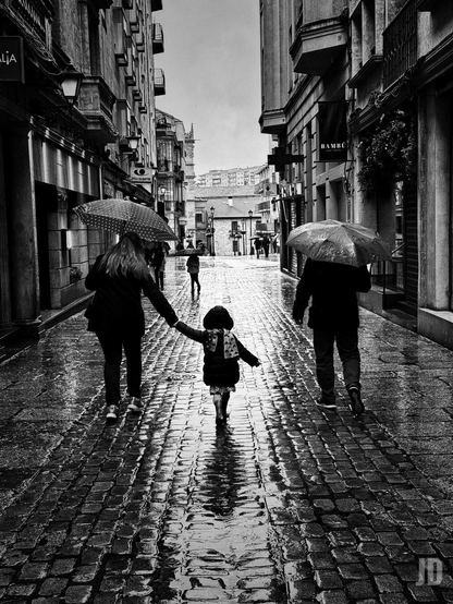 Esta es una evocadora fotografía en blanco y negro capturada en la Calle Prior de Salamanca. La imagen transmite una atmósfera melancólica y serena propia de un día lluvioso en una ciudad histórica.
• Composición Central: En el primer plano, vemos a una familia caminando de espaldas. Una mujer a la izquierda y un hombre a la derecha llevan paraguas, mientras que una niña pequeña camina en medio, de la mano de la mujer, vistiendo un abrigo oscuro y una bufanda estampada.
• Texturas y Reflejos: El suelo de pavimento de piedra mojado es el gran protagonista técnico de la foto. El agua crea reflejos verticales que alargan las siluetas de los peatones, aportando profundidad y brillo a la escena.
• Entorno Urbano: Los edificios de piedra de Villamayor, típicos de Salamanca, flanquean la calle estrecha. Se pueden apreciar carteles comerciales (como el de "Bambú"), farolas clásicas de pared y balcones con herrajes metálicos.
El uso del alto contraste resalta los detalles de las piedras y el brillo del agua, creando una sensación de atemporalidad. La perspectiva conduce la mirada hacia el fondo de la calle, donde se vislumbran más figuras bajo paraguas y la arquitectura característica de la zona monumental. Es una estampa clásica que captura un momento cotidiano cargado de nostalgia.