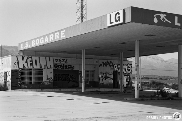 Abandoned petrol station with a weathered structure, featuring large signs and walls covered in graffiti. The surrounding landscape includes mountains in the background and an empty parking area, depicted in black and white.