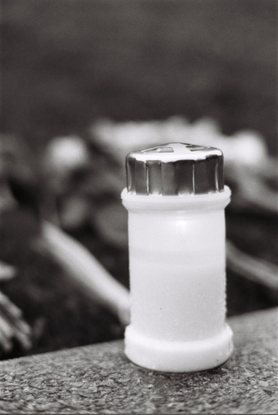 Black-and-white photograph of a white grave lantern sitting on a stone ledge. In the blurry background lie several bouquets of flowers, barely recognisable.