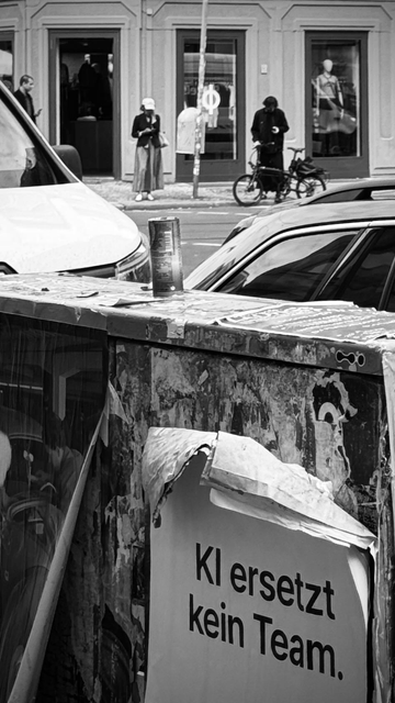 A black-and-white street scene featuring a woman in a cap and skirt looking at her phone, standing next to a storefront. In the foreground, there is a graffiti-covered surface with a partially torn poster that reads "KI ersetzt kein Team"